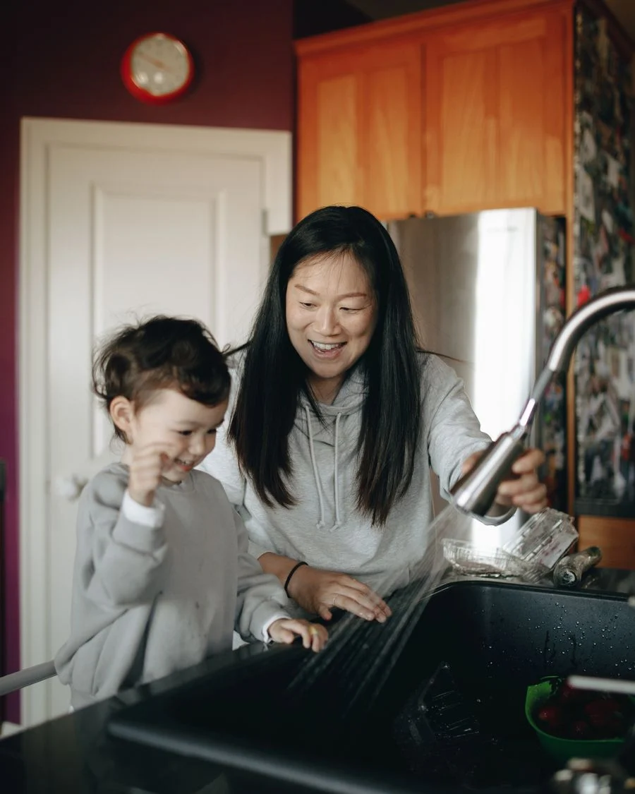 A mother and child happily washing hands together at the kitchen sink